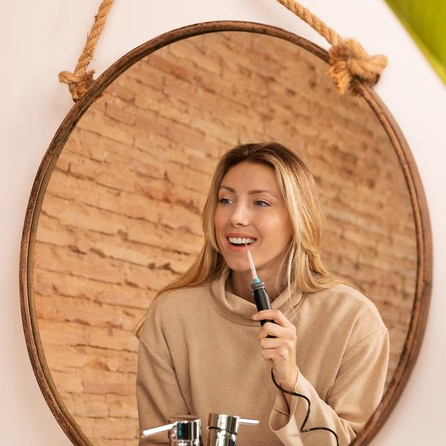 Kobieta myjąca zęby przed lustrem. Uśmiecha się i trzyma szczoteczkę.