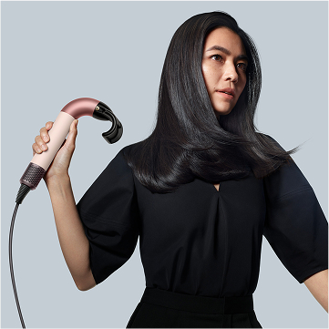 Una mujer con cabello oscuro sostiene un secador de pelo rosa dorado y beige. El fondo es gris.