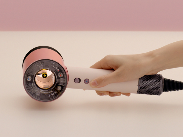 Una mano sosteniendo un secador de pelo rosa dorado y blanco, primer plano contra un fondo rosa.