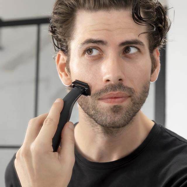 Mężczyzna goli policzek czarną trymerem. Patrzy w lewo. Białe tło.