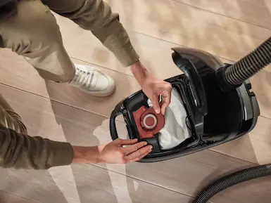 Bir kişi, ahşap bir zeminde bir filtre çıkararak elektrik süpürgesini boşaltıyor.