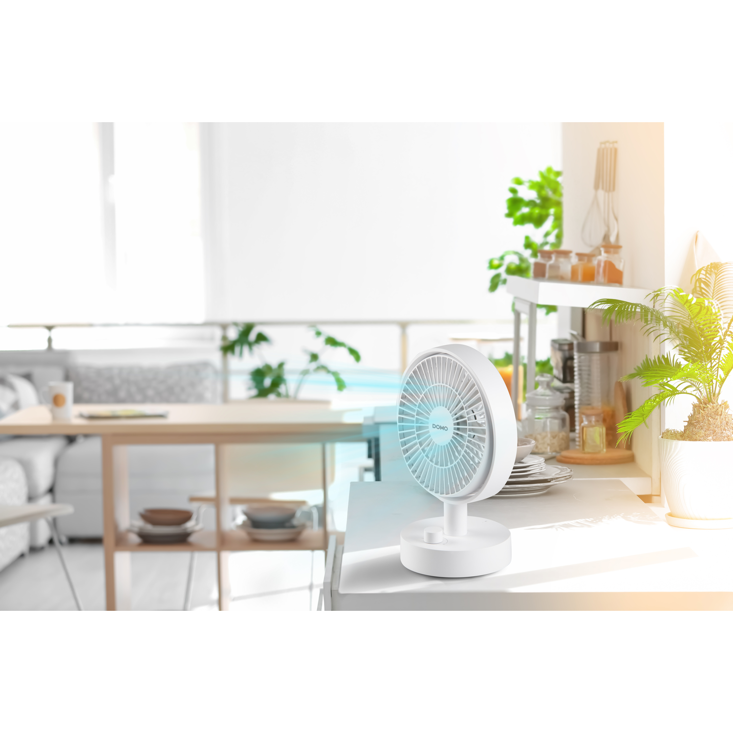 A white desk fan on a countertop in a bright room with a window and plants. It is turned on.