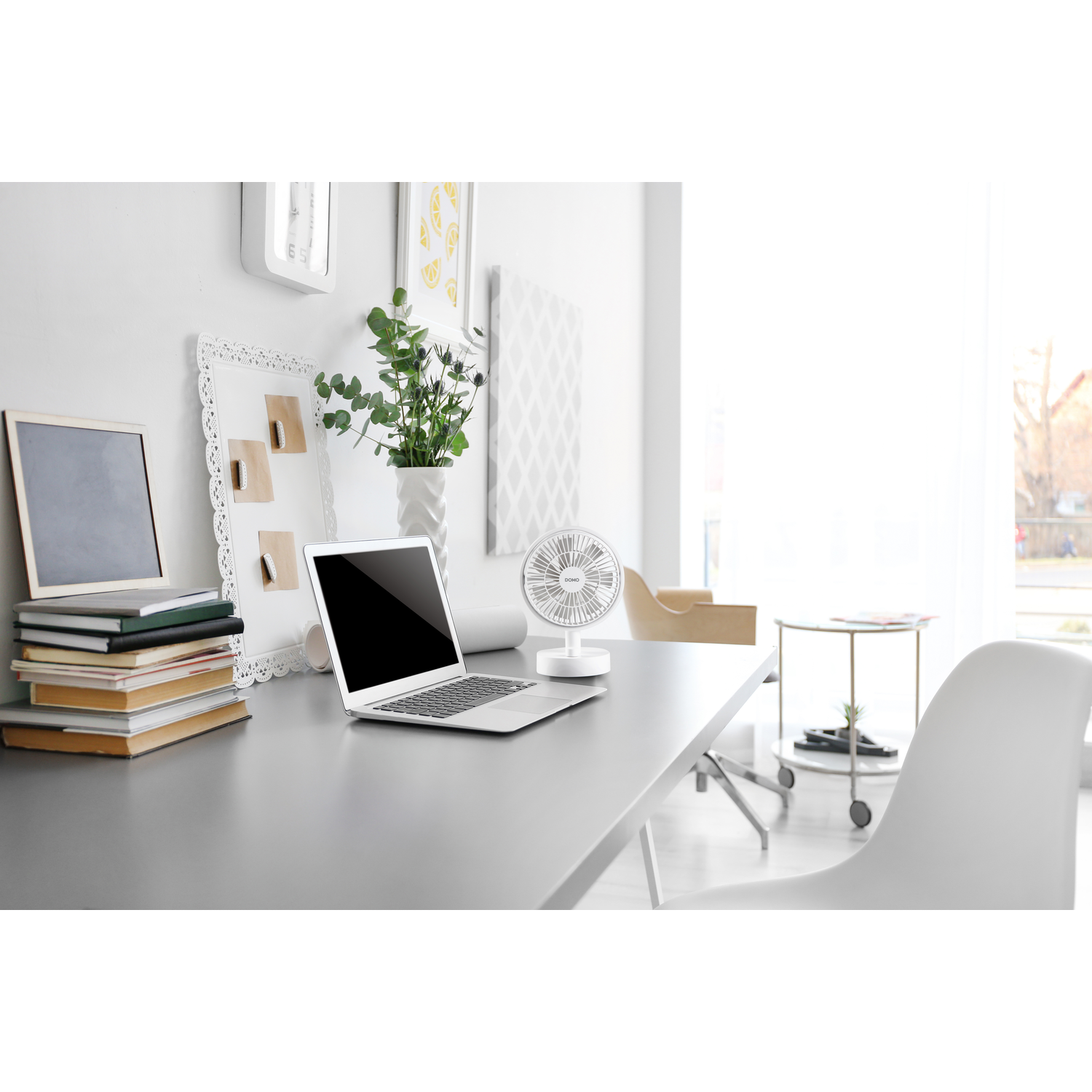 A desk with a laptop and a small fan. There are books and decorations. The room is bright.