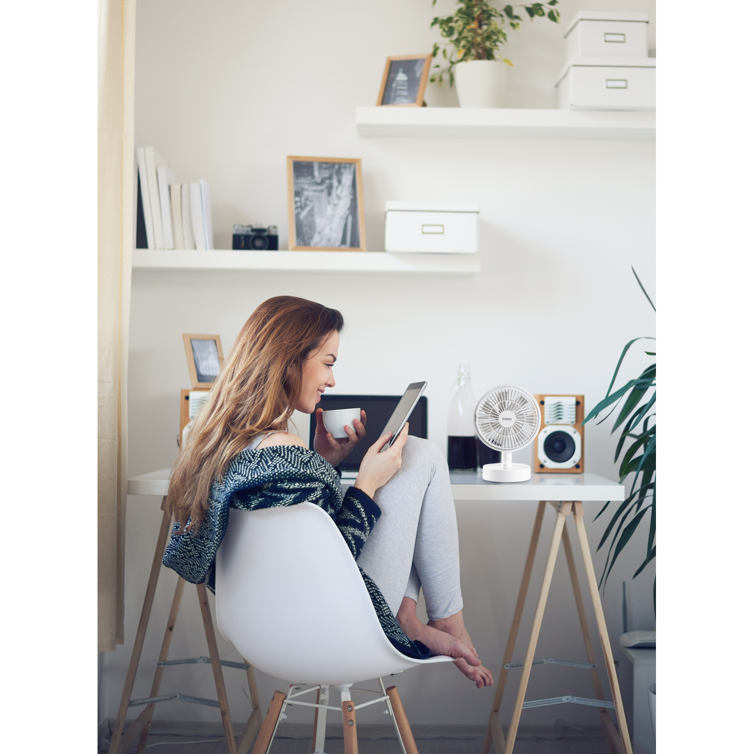 Woman using a tablet, sitting at a desk with a fan, speakers, and a window behind her.