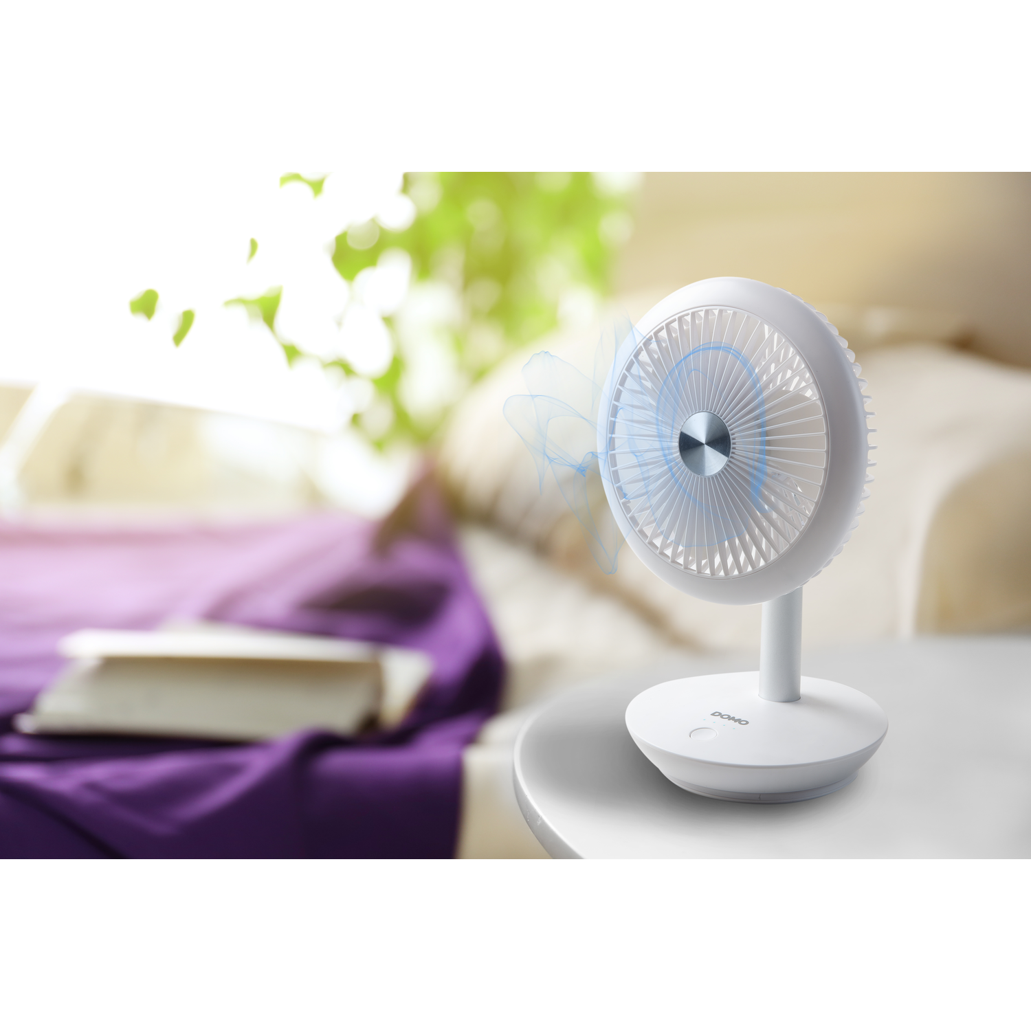 White desk fan with a blue blade effect, on a table next to a bed and books.