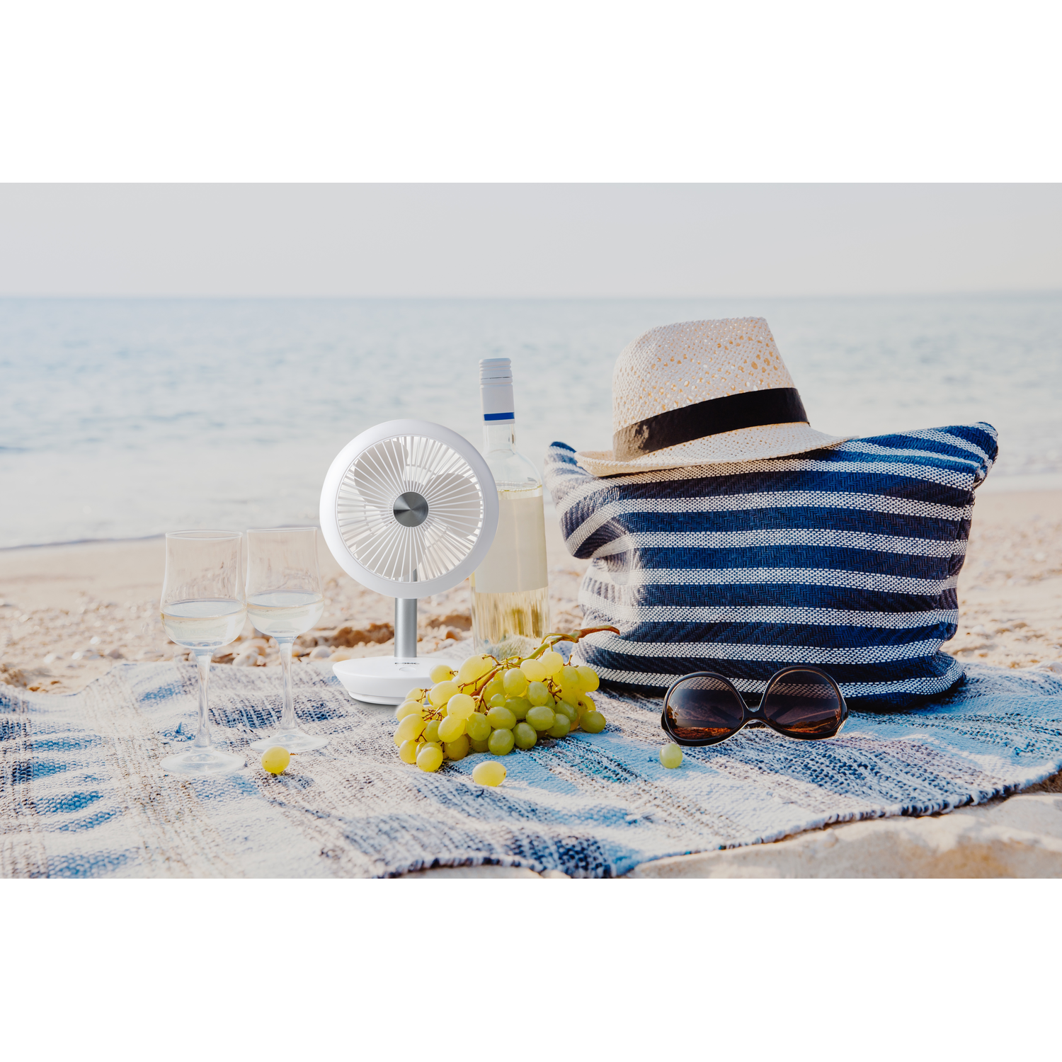 A picnic on a sandy beach with a fan, wine, hat, sunglasses, and grapes.