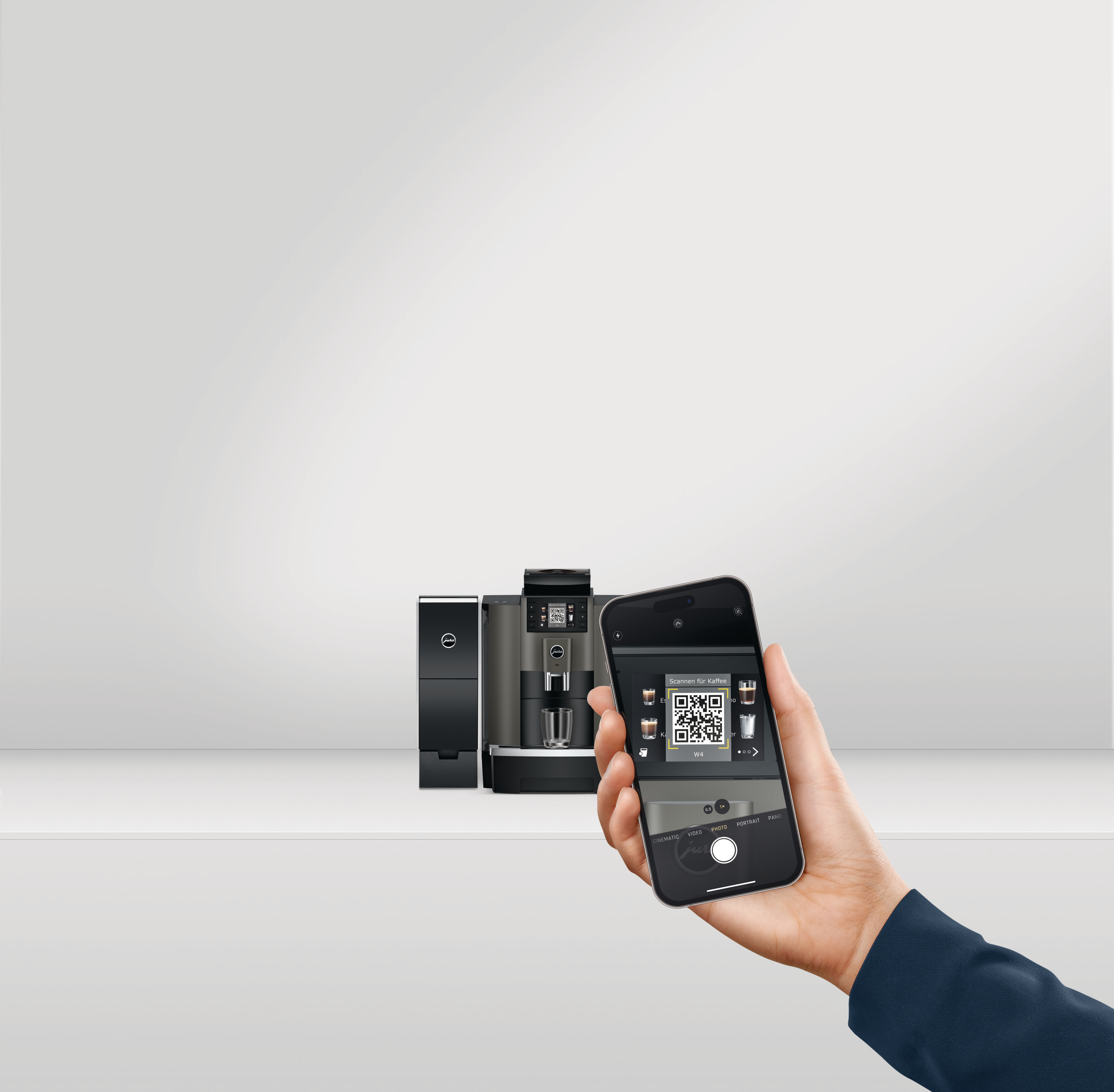 Hand hält Telefon über Espressomaschine. Das Telefon zeigt einen QR-Code.