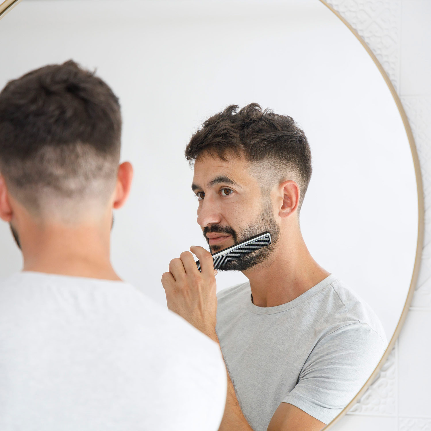 Hombre en espejo, peinándose la barba frente a una pared blanca.