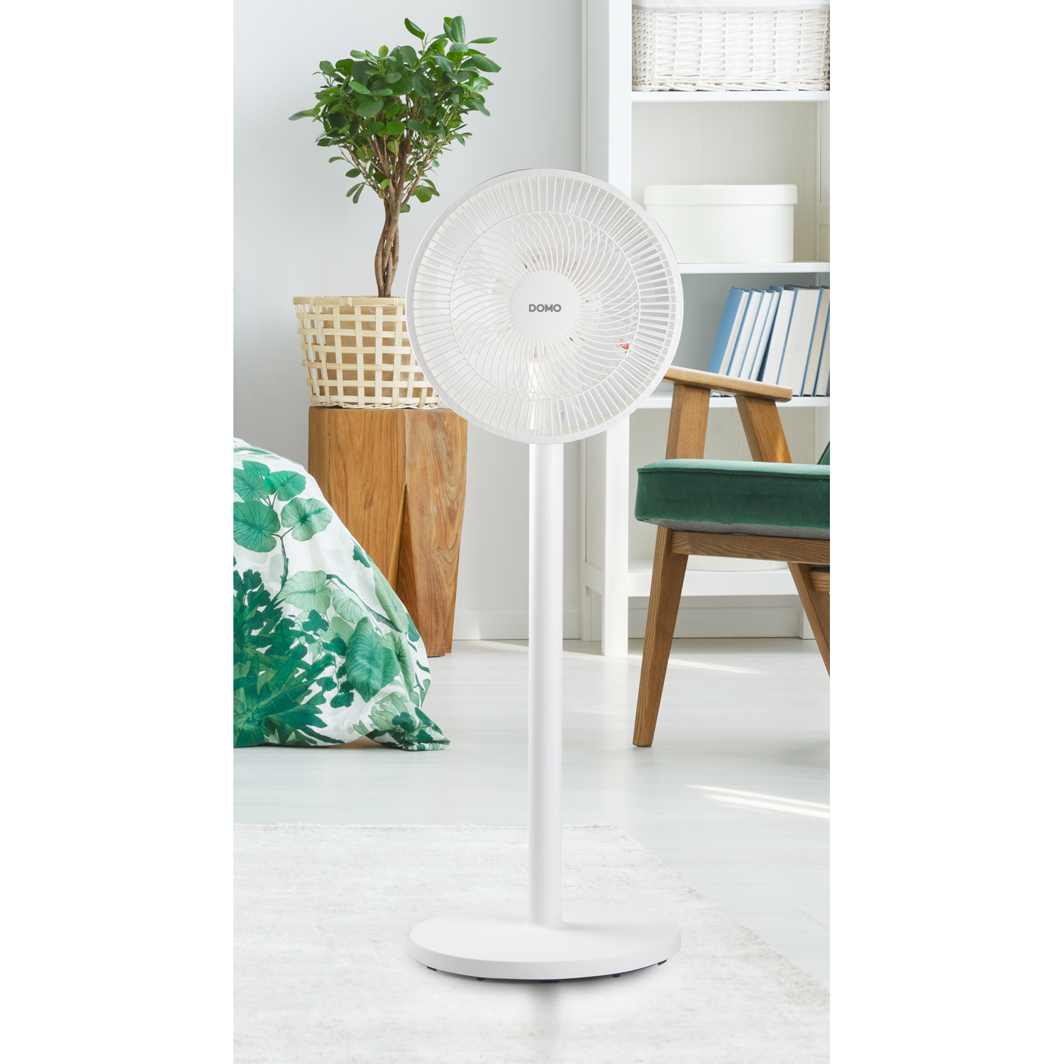 Ventilateur sur pied blanc dans une pièce. Une couverture à imprimé de feuilles vertes est visible sur un lit.