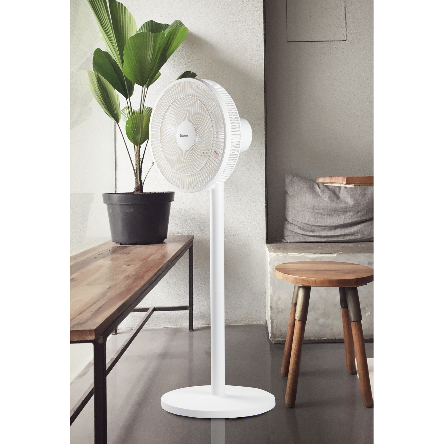 Ventilateur sur pied blanc dans une pièce avec une plante, un banc et un tabouret en bois.