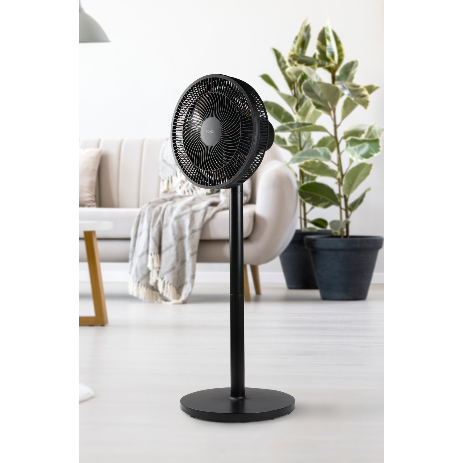 Ventilateur sur pied noir dans une pièce. Un canapé et des plantes en arrière-plan.