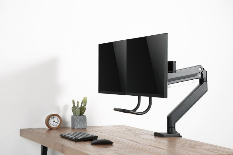 Kettős számítógépes monitorok egy asztalon, állítható karra szerelve, kaktusszal és órával.