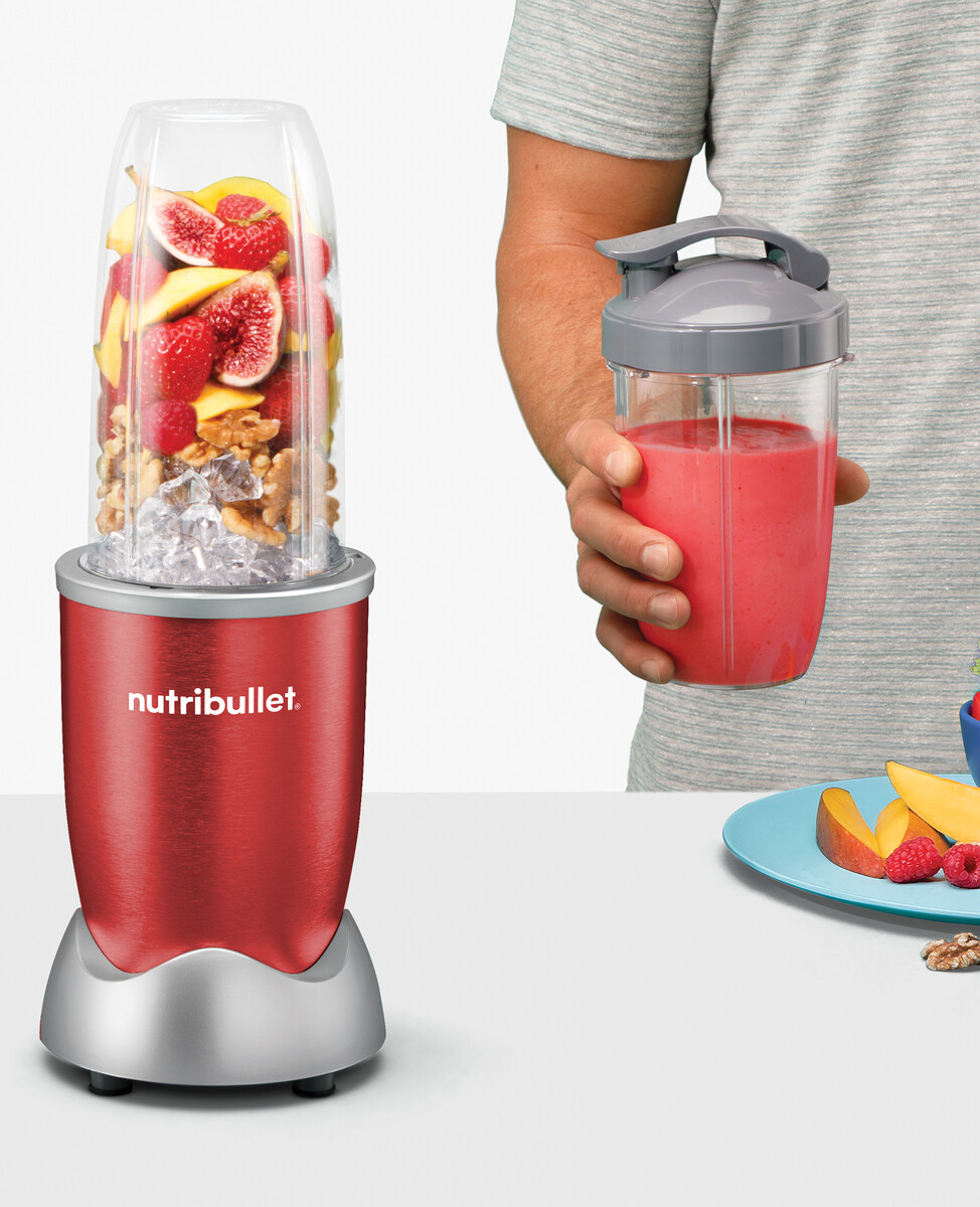 Czerwony blender Nutribullet ze składnikami w środku i koktajlem w kubku w dłoni.