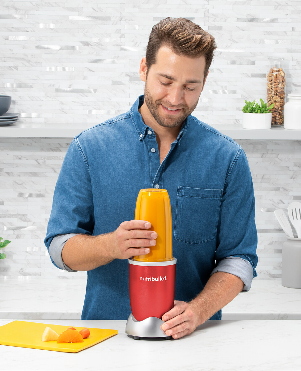 Mężczyzna w niebieskiej koszuli dodaje płyn do czerwonego blendera, składniki z boku.