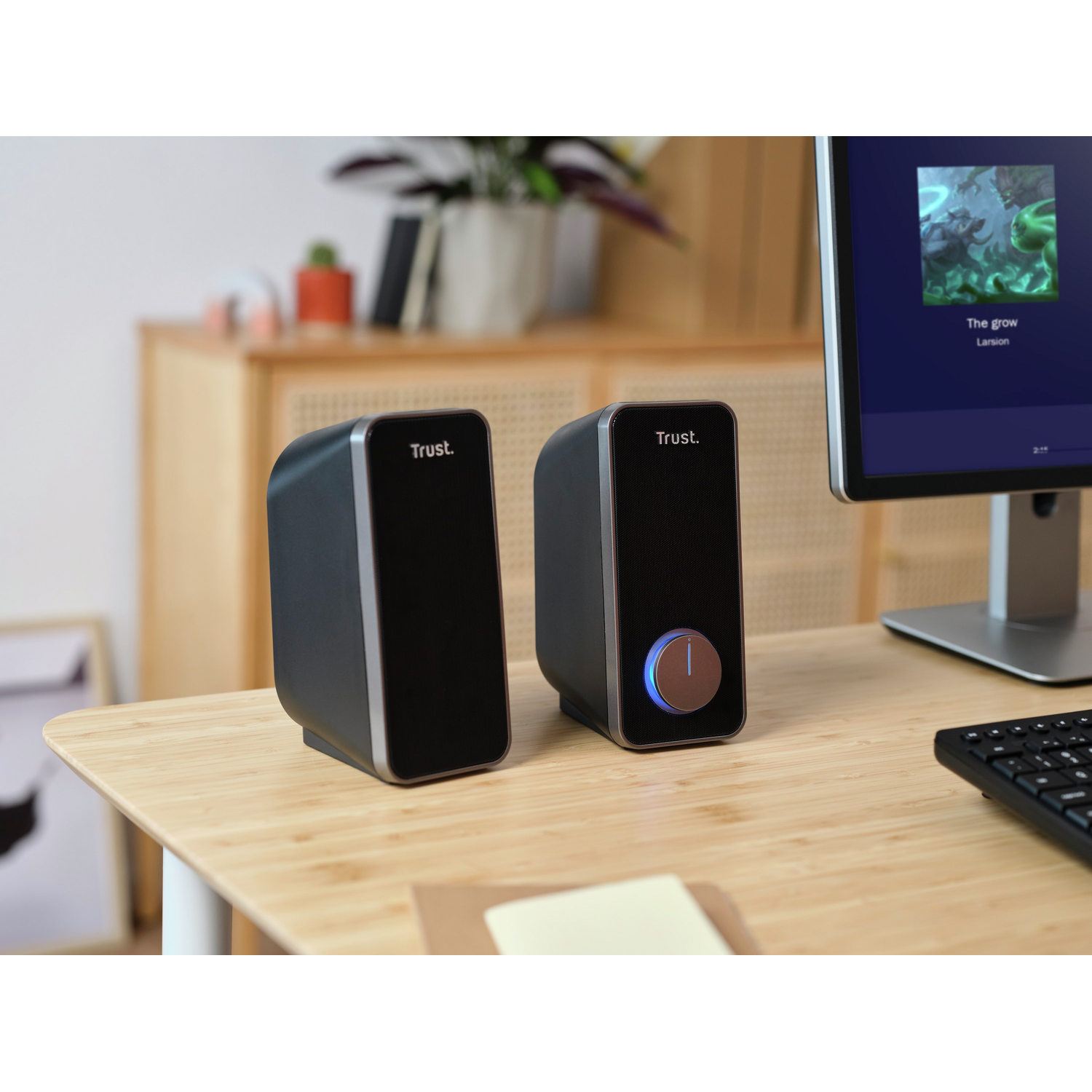 Two black speakers on a wooden table with a computer monitor and a keyboard visible in the background.