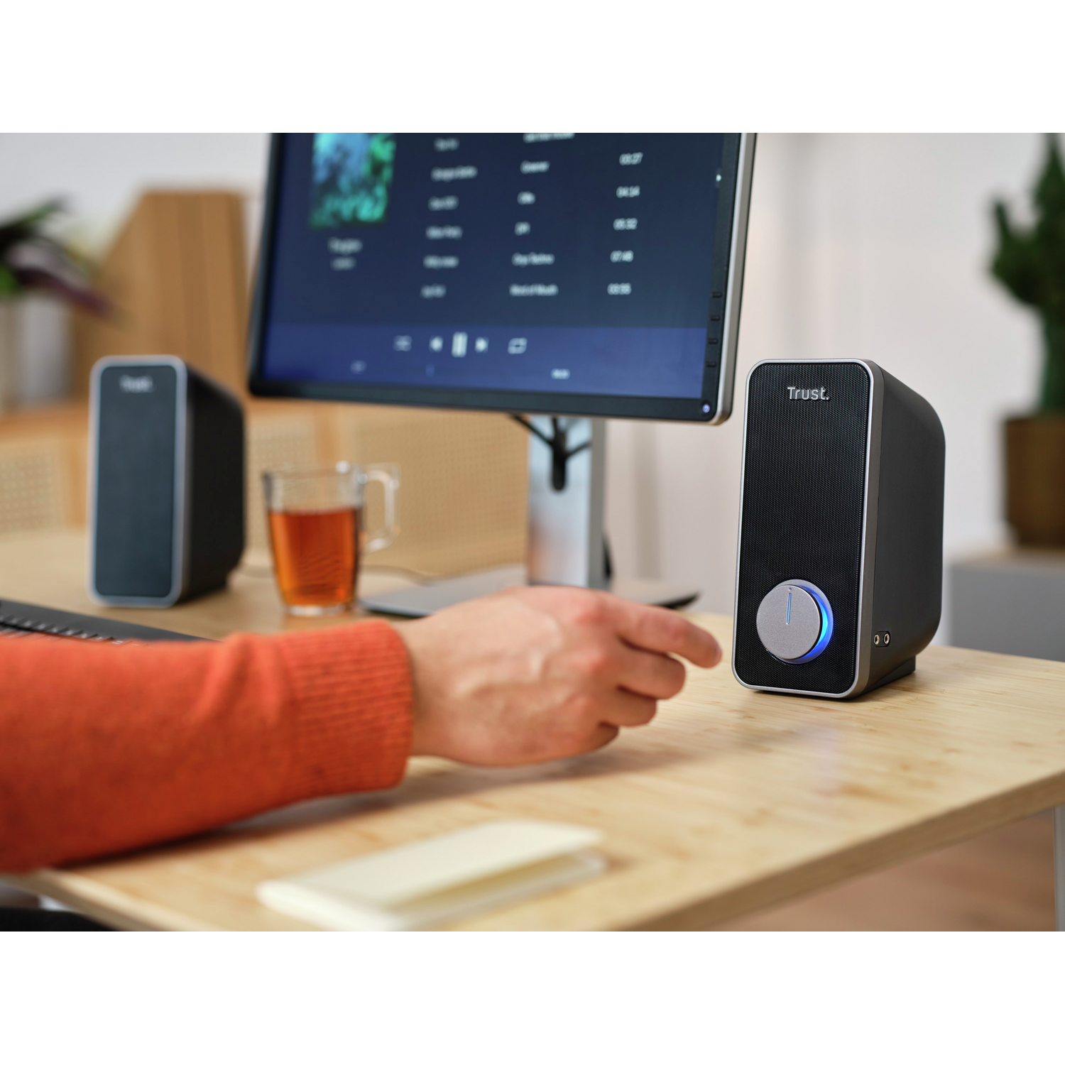 Person's hand near a speaker, with a monitor displaying music. Wooden desk.
