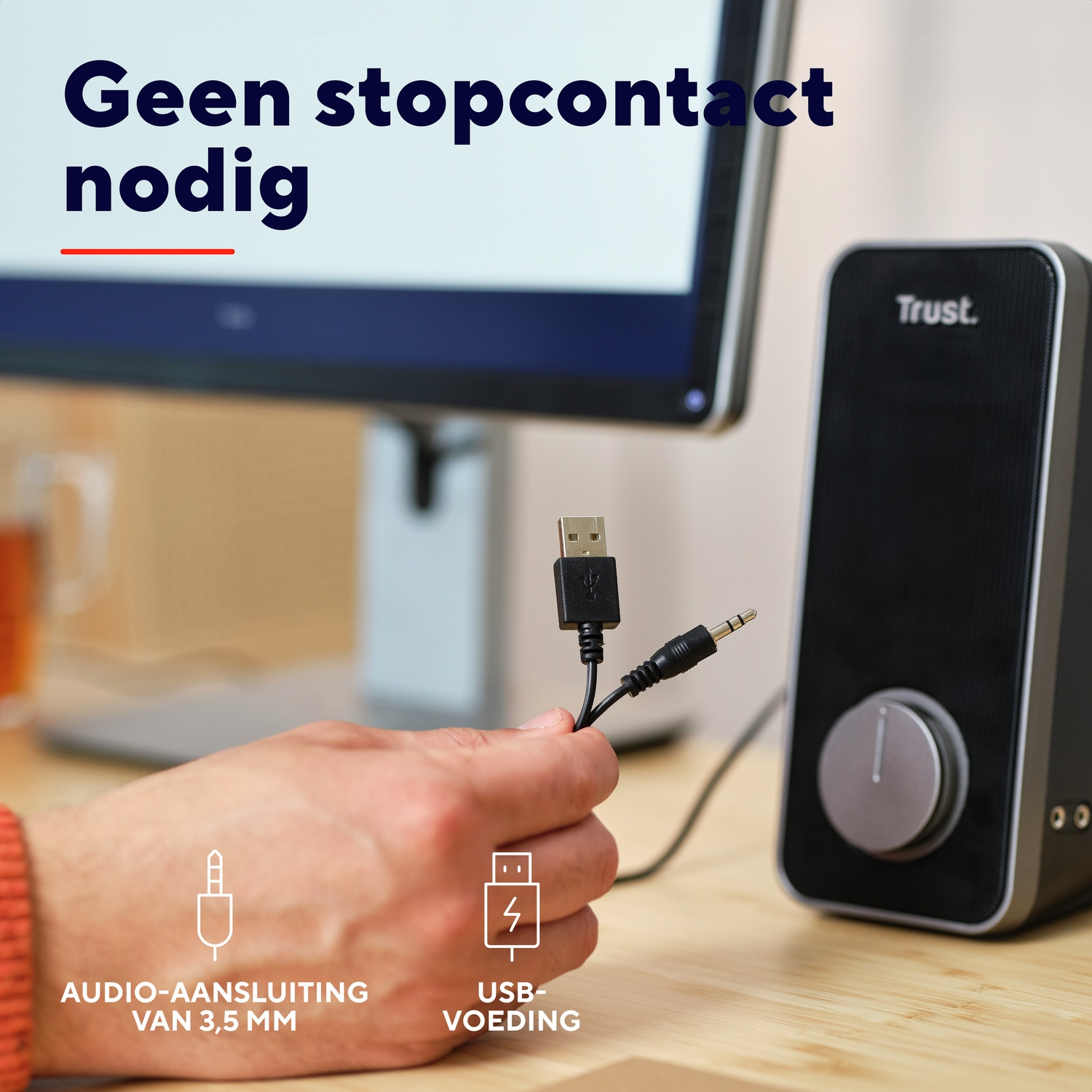 Een hand die een USB en een 3,5 mm jackkabel vasthoudt naast een Trust-luidspreker. De luidspreker is zwart.