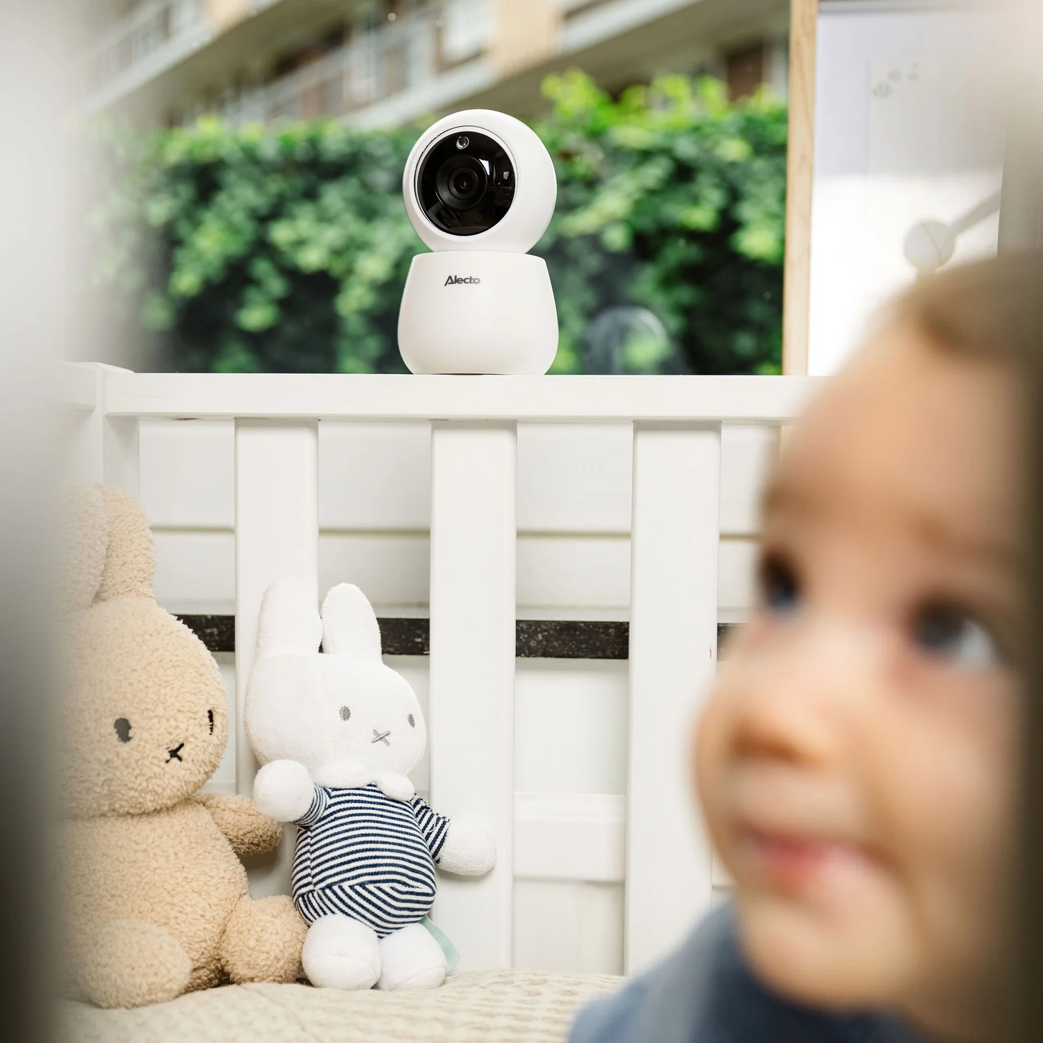 A baby monitor in a crib with a baby looking at it. Stuffed animals nearby.