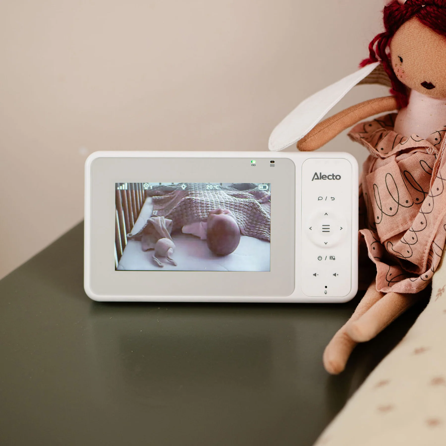 Baby monitor showing a baby in a crib. A doll is next to the monitor. Soft background.