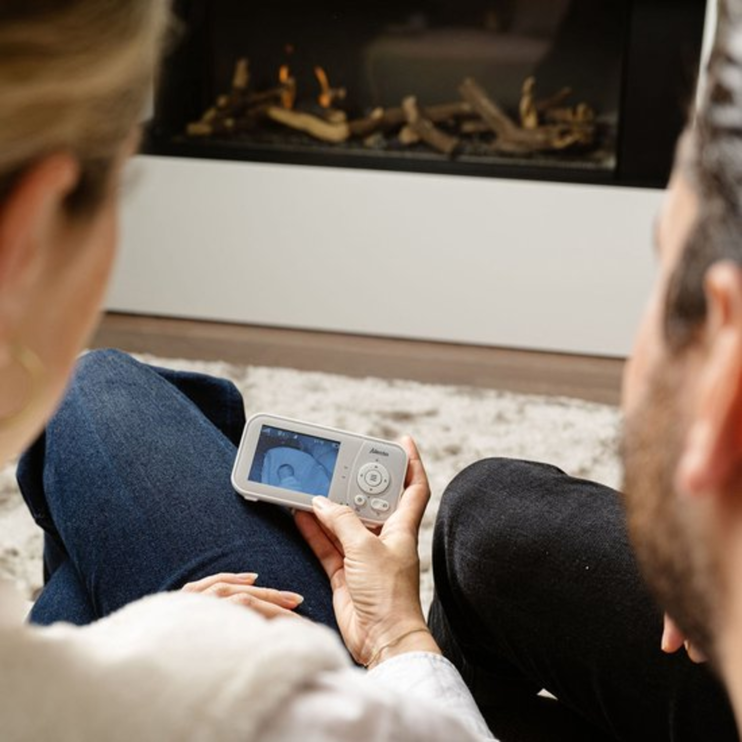 A couple watches a baby monitor. A fireplace is in the background.