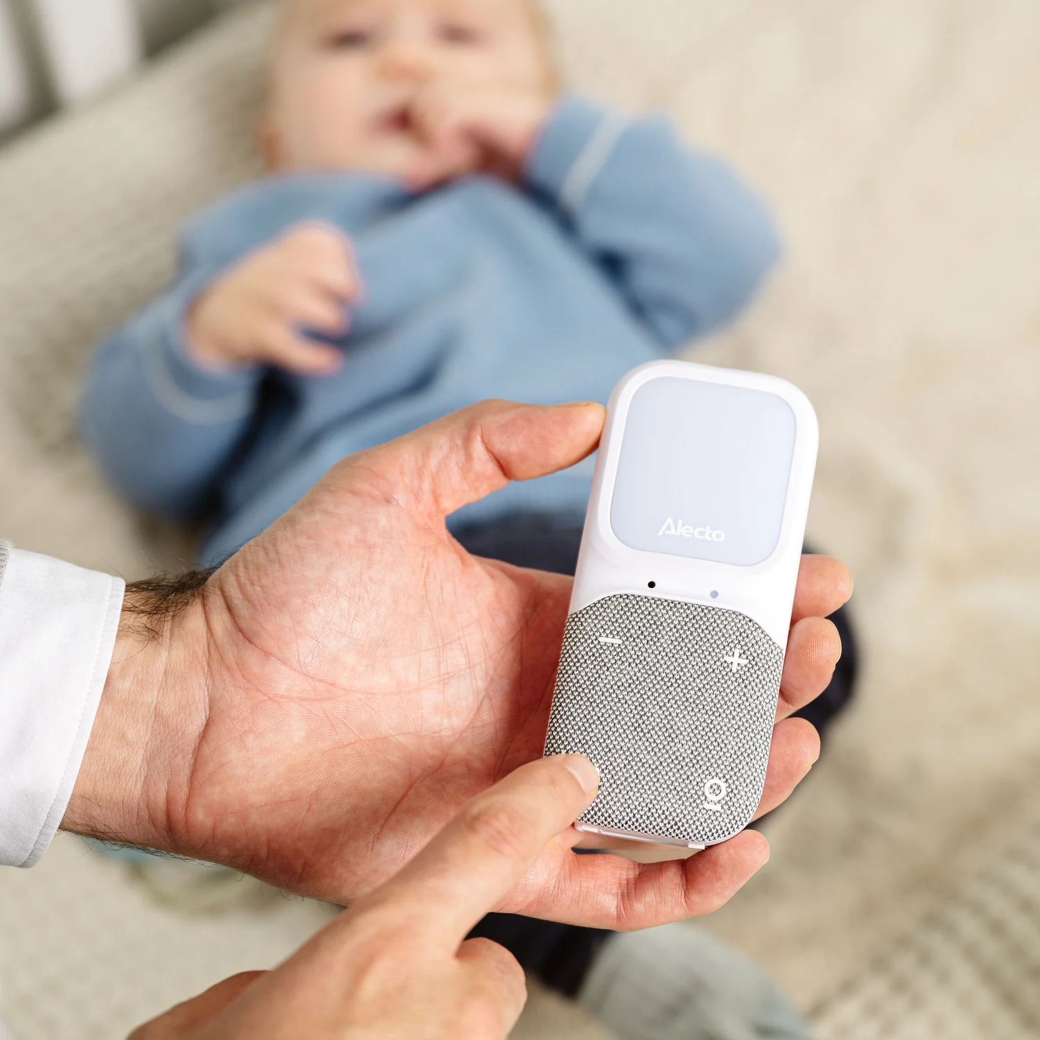 A hand holding a white and gray baby monitor, with a baby blurred in the background.