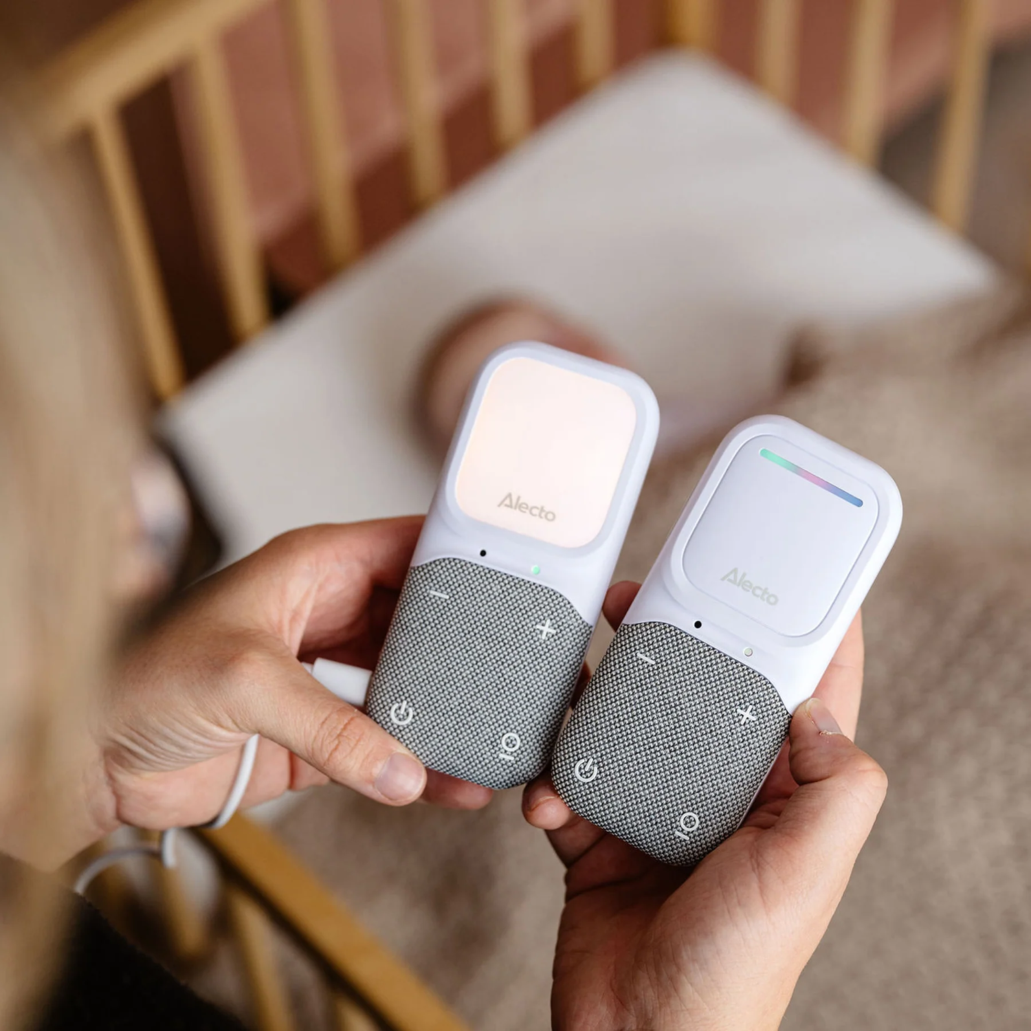 Two white baby monitors. Hands holding them in front of a blurred crib background.