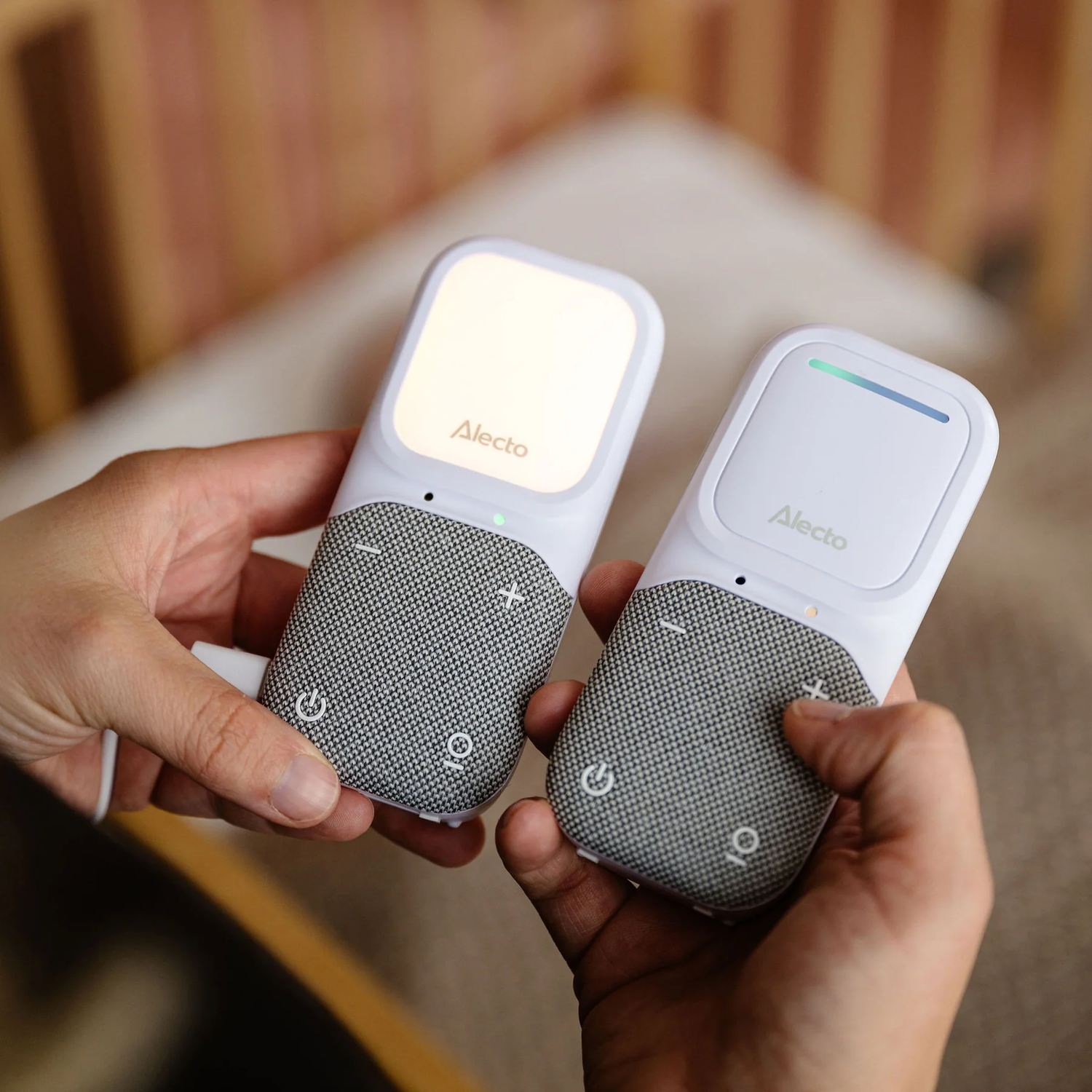 Two white baby monitors, one on each hand. They are white with gray mesh and the Alecto brand.