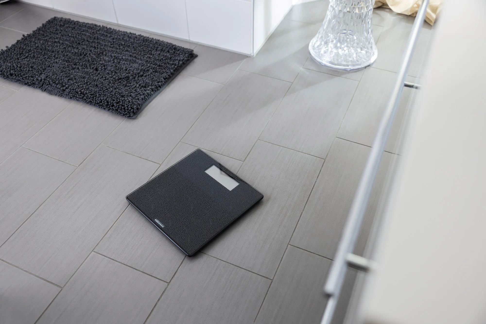 Bilancia digitale nera su un pavimento piastrellato in un bagno con un tappetino.
