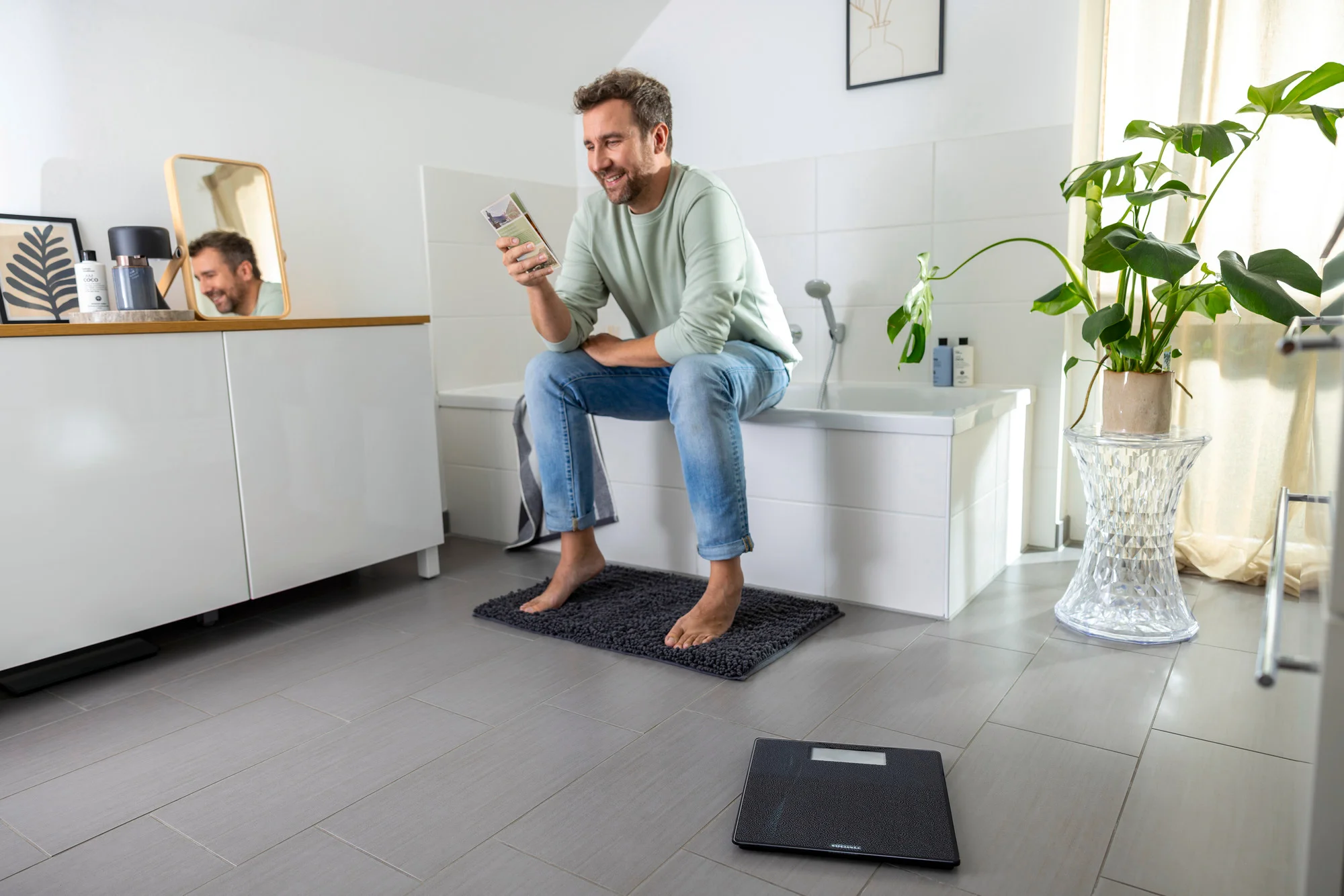Uomo in bagno guarda il telefono, bilancia sul pavimento. Bianco, grigio, legno.