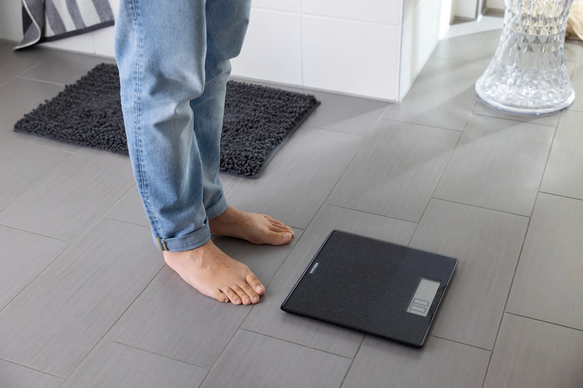Persona in piedi su un pavimento piastrellato grigio, che guarda una bilancia digitale nera. C'è un tappetino da bagno nelle vicinanze.
