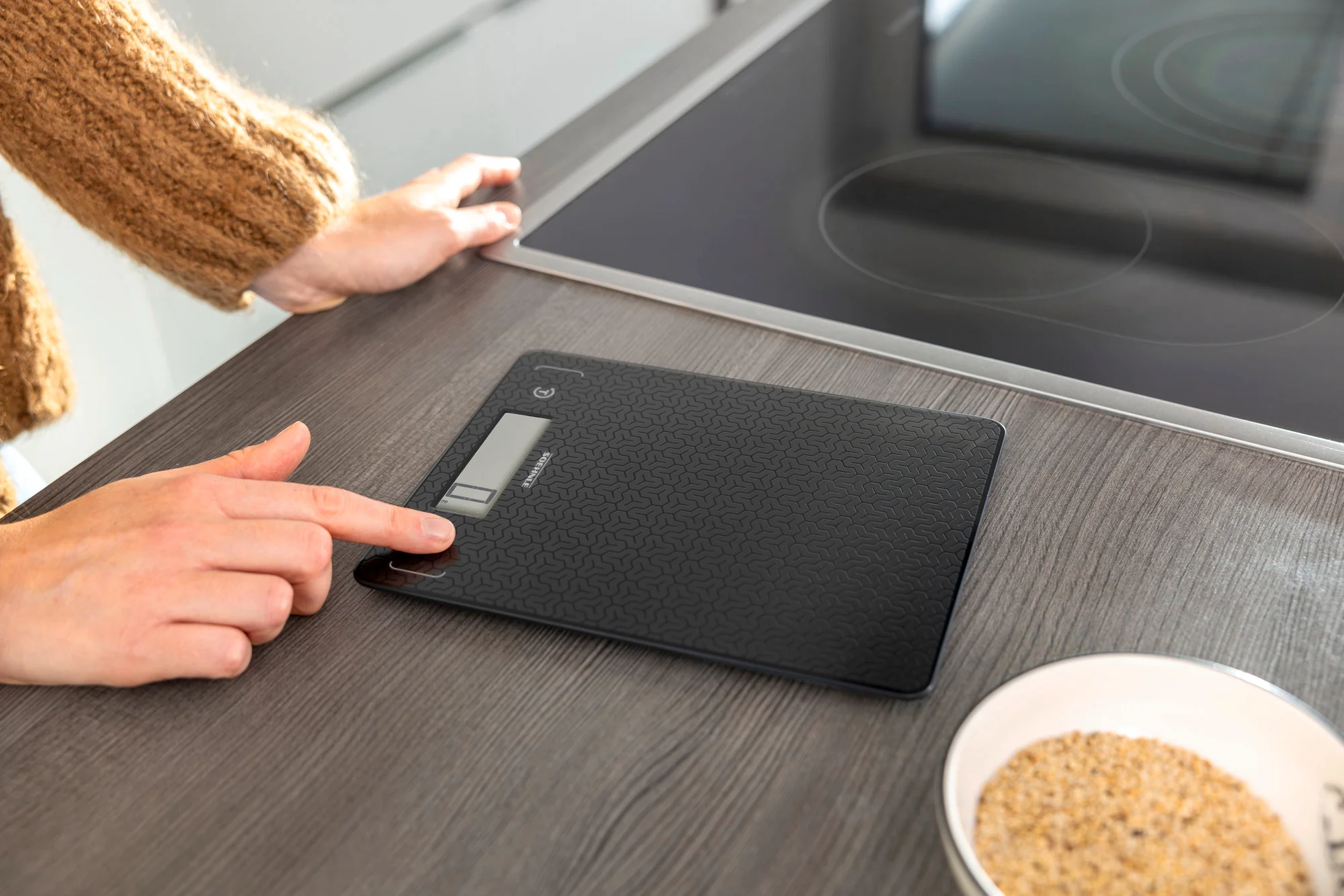 Balance de cuisine numérique noire sur une surface en bois. Une main utilise la balance.