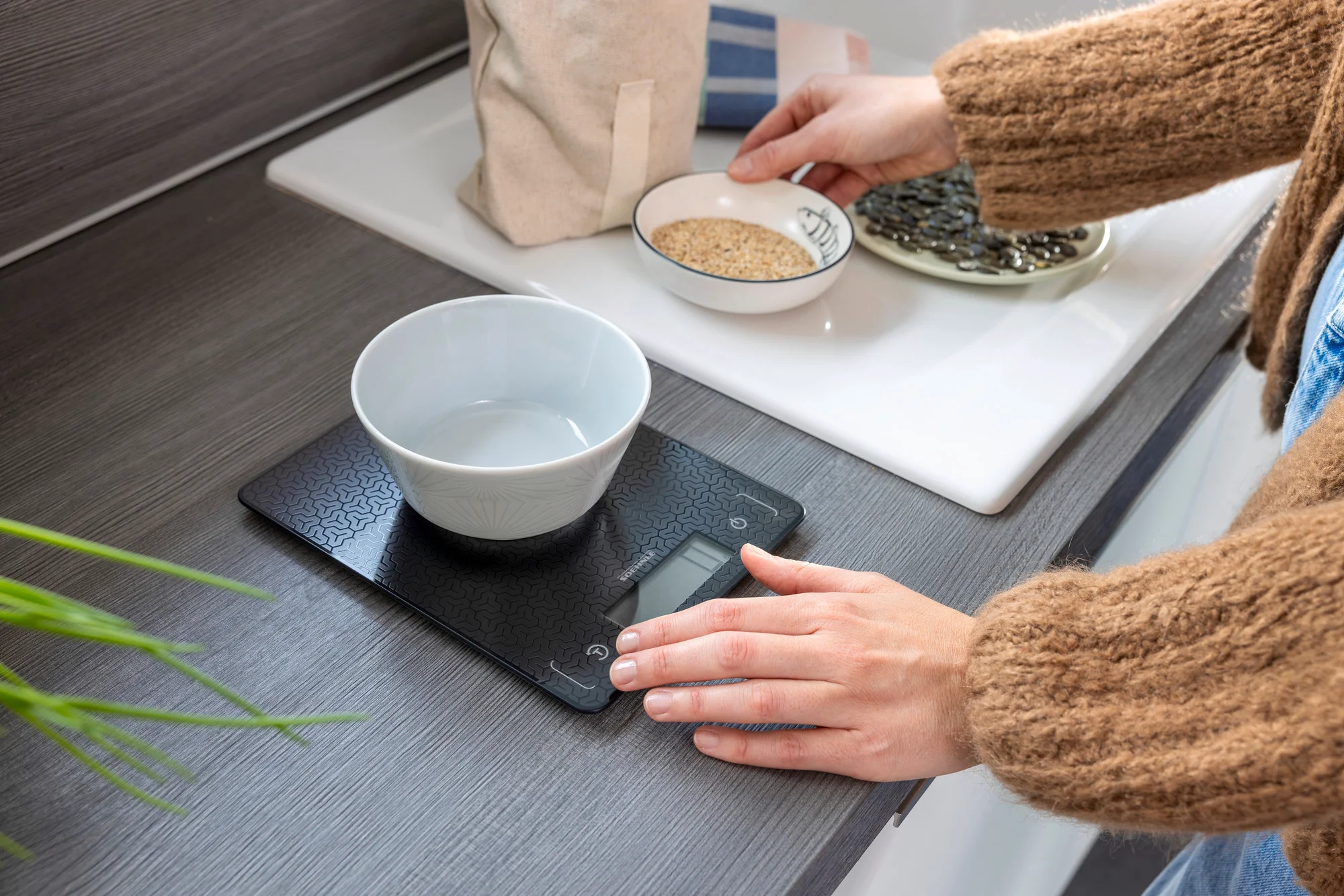 Mains sur une balance numérique avec un bol. Autres bols avec des graines. Plan de travail de cuisine.
