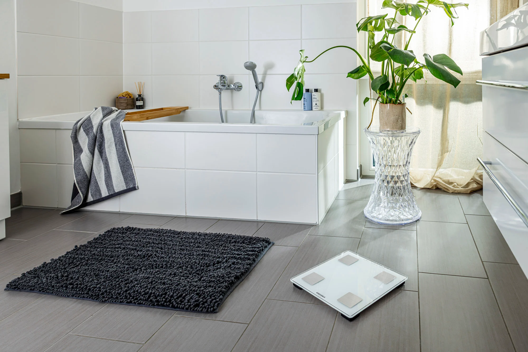 Scène de salle de bain avec baignoire, balance et plante sur tabouret transparent.