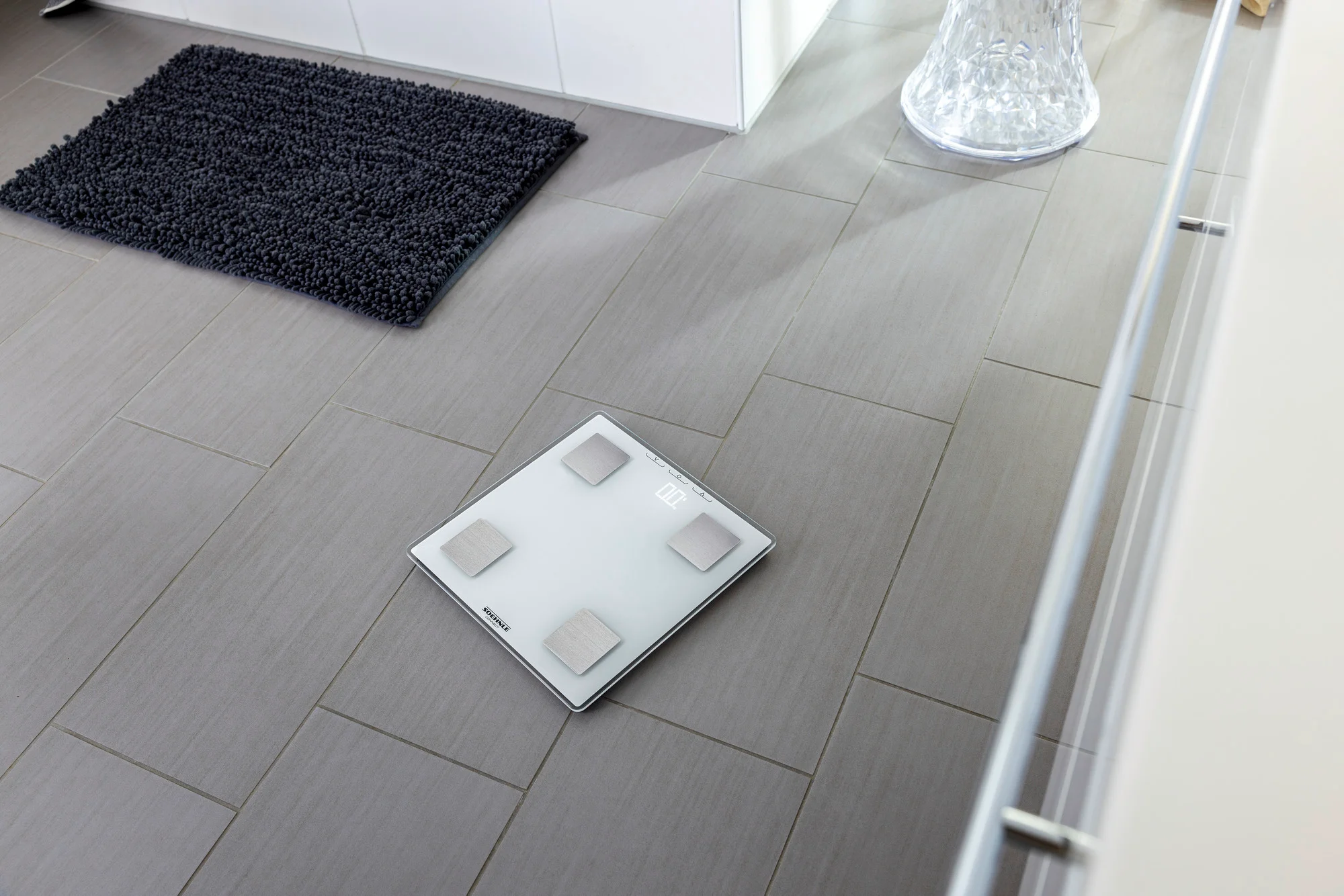 Une balance blanche sur un sol carrelé gris. Un tapis noir est à l'arrière-plan.