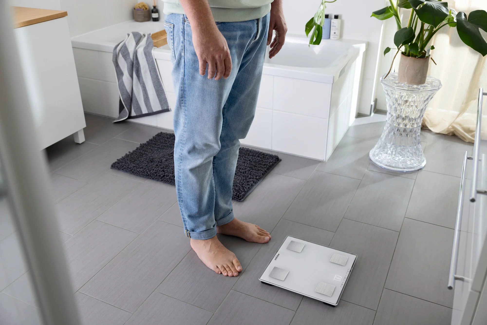 Un homme en jean est debout sur une balance dans une salle de bain. Une serviette et une plante sont en arrière-plan.