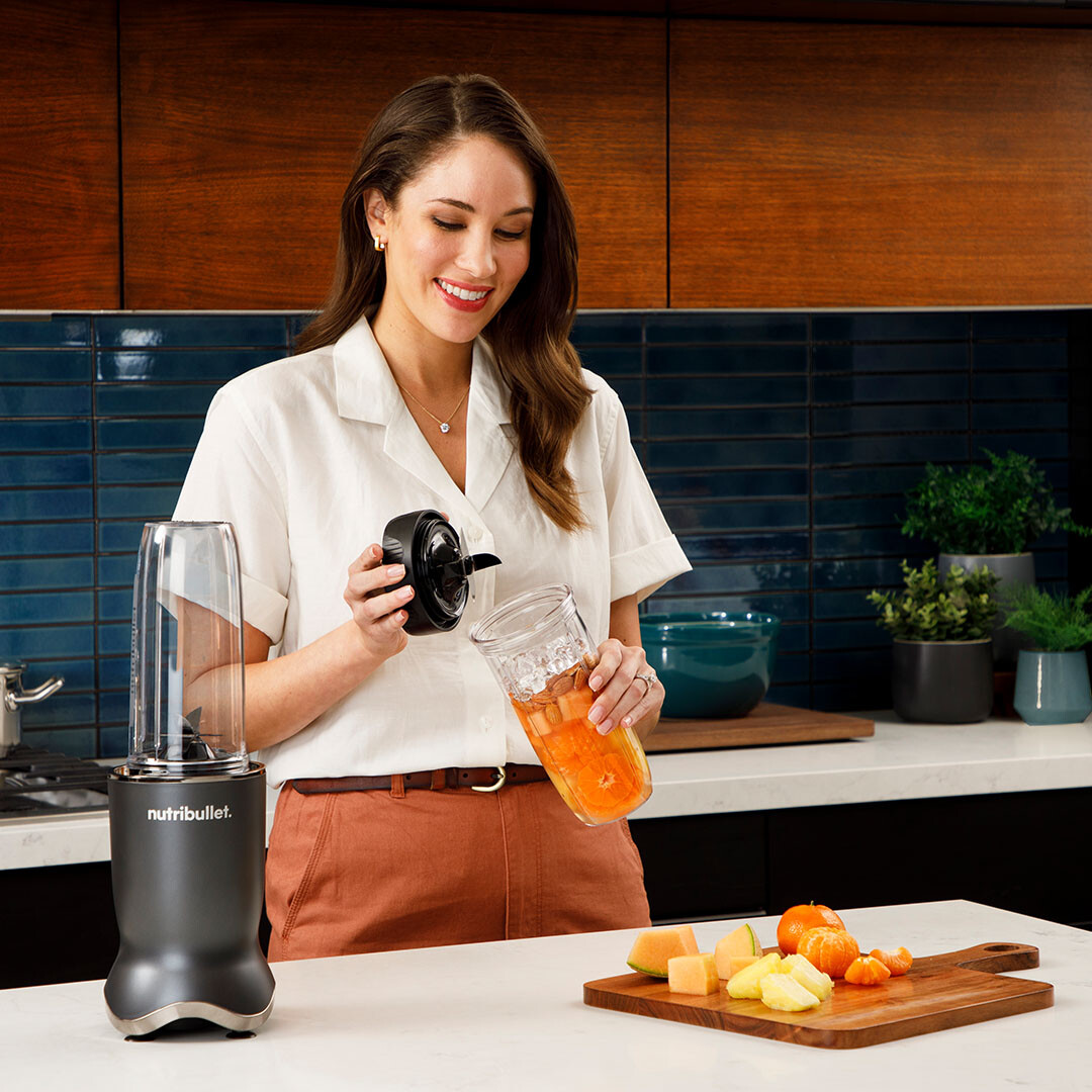 Kobieta z blenderem Nutribullet. Uśmiecha się i trzyma pokrywę blendera. Biały blat.