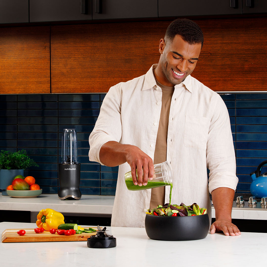 Mężczyzna wlewa zielone smoothie do miski z sałatką. Blender i składniki na stole.