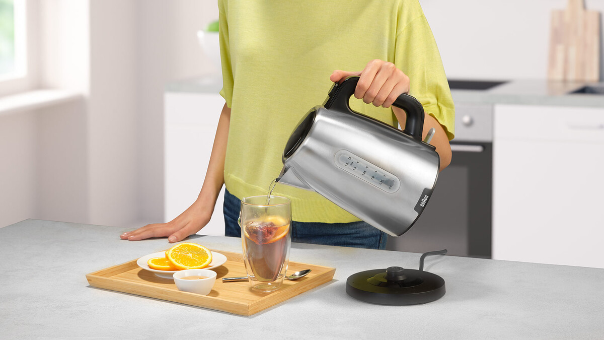 Osoba wlewa wodę ze srebrnego czajnika do szklanki z herbatą i plasterkami pomarańczy. Na stole taca z plasterkami pomarańczy i małą miseczką.