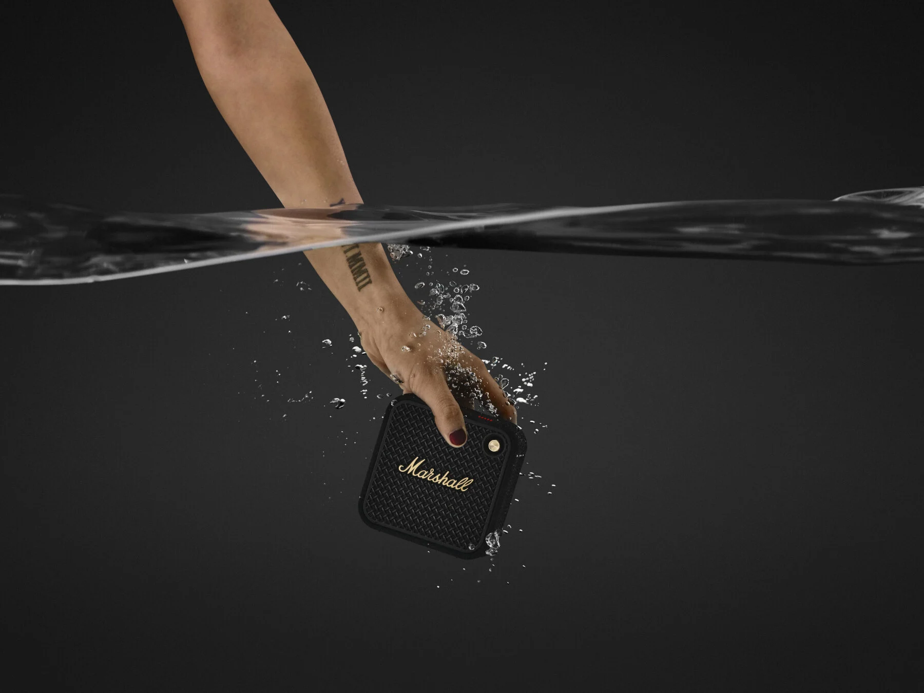 Mano che immerge un altoparlante nero nell'acqua. Schizzi d'acqua. Logo Marshall visibile.