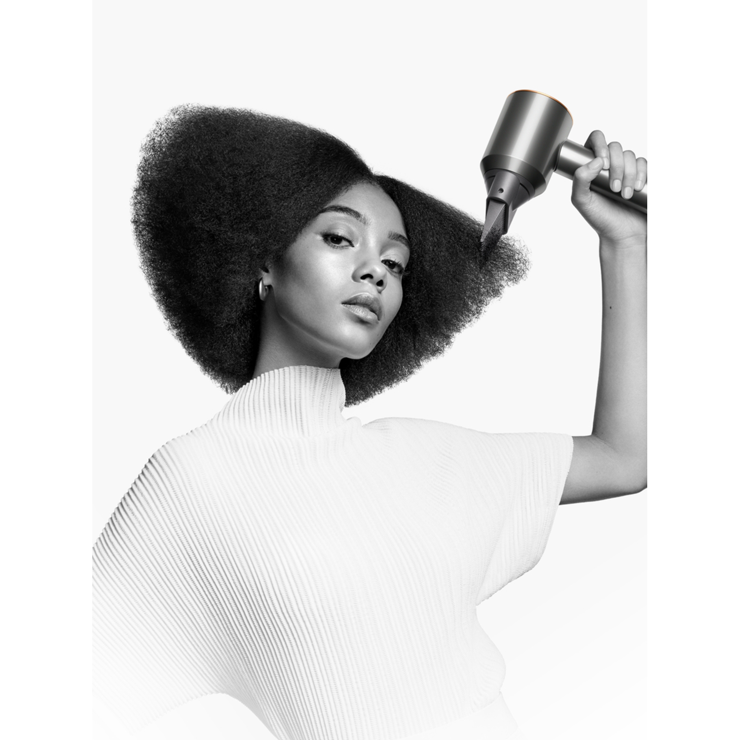 A woman with big hair is drying her hair with a silver hair dryer.