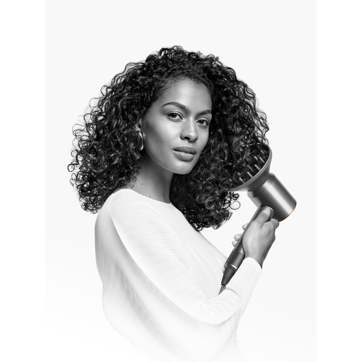 Woman holding a silver hair dryer. She is looking at the camera, with curly hair and white background.