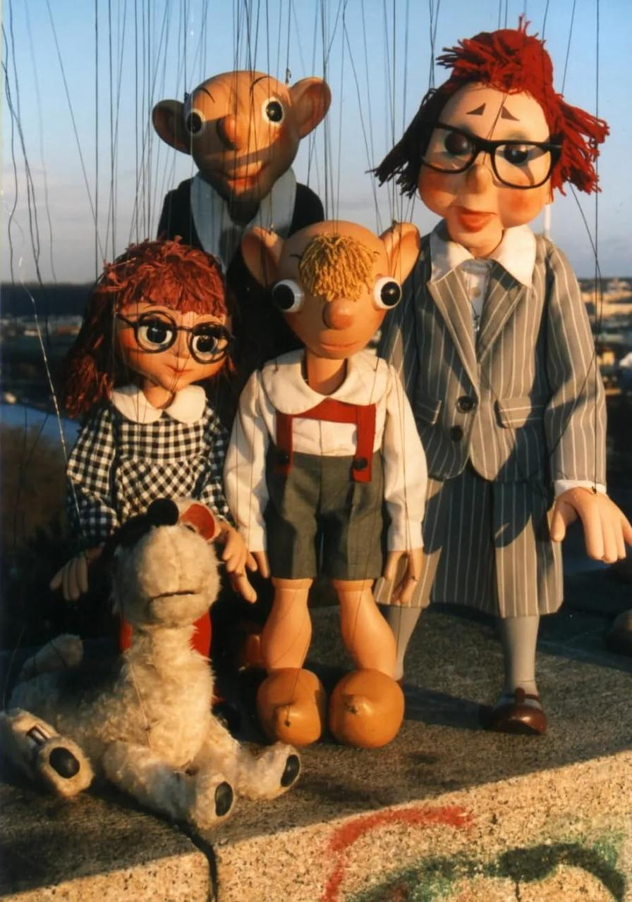 Puppen auf einer Bühne. Fünf Puppen sind sichtbar, mit einer Stadtkulisse.