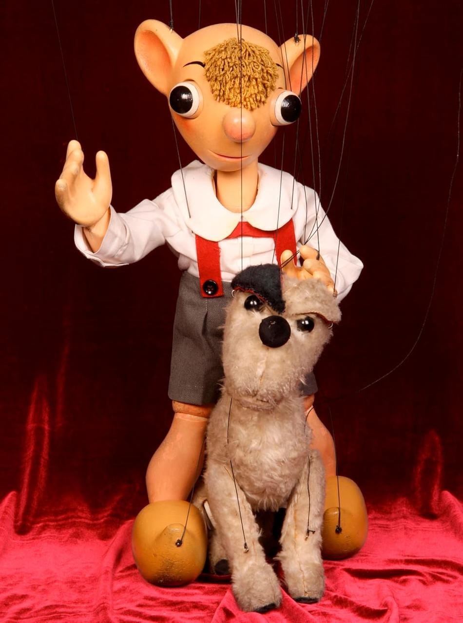 Eine Marionette mit großem Kopf und kleinem Körper hält eine kleinere Marionette eines Hundes. Der Hintergrund ist dunkelrot.