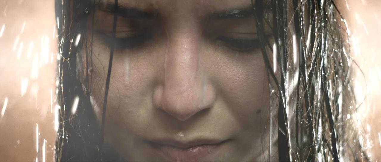 Eine Frau mit nassem Haar, geschlossenen Augen und Wassertropfen. Dunkler Hintergrund mit unscharfen Lichtern.