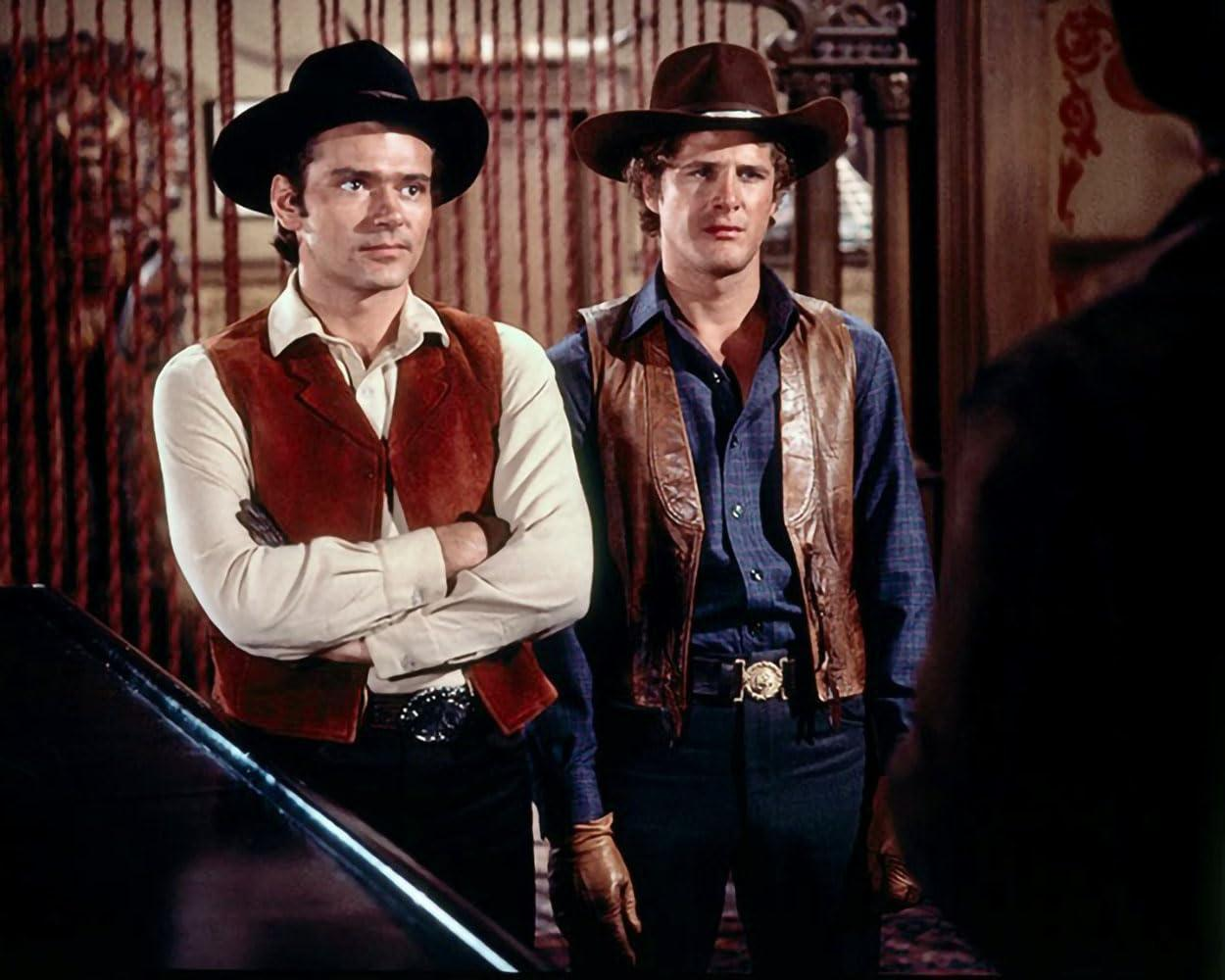 Zwei Männer in Cowboy-Kleidung schauen nach vorne, braune und schwarze Hüte, stehen im Raum.
