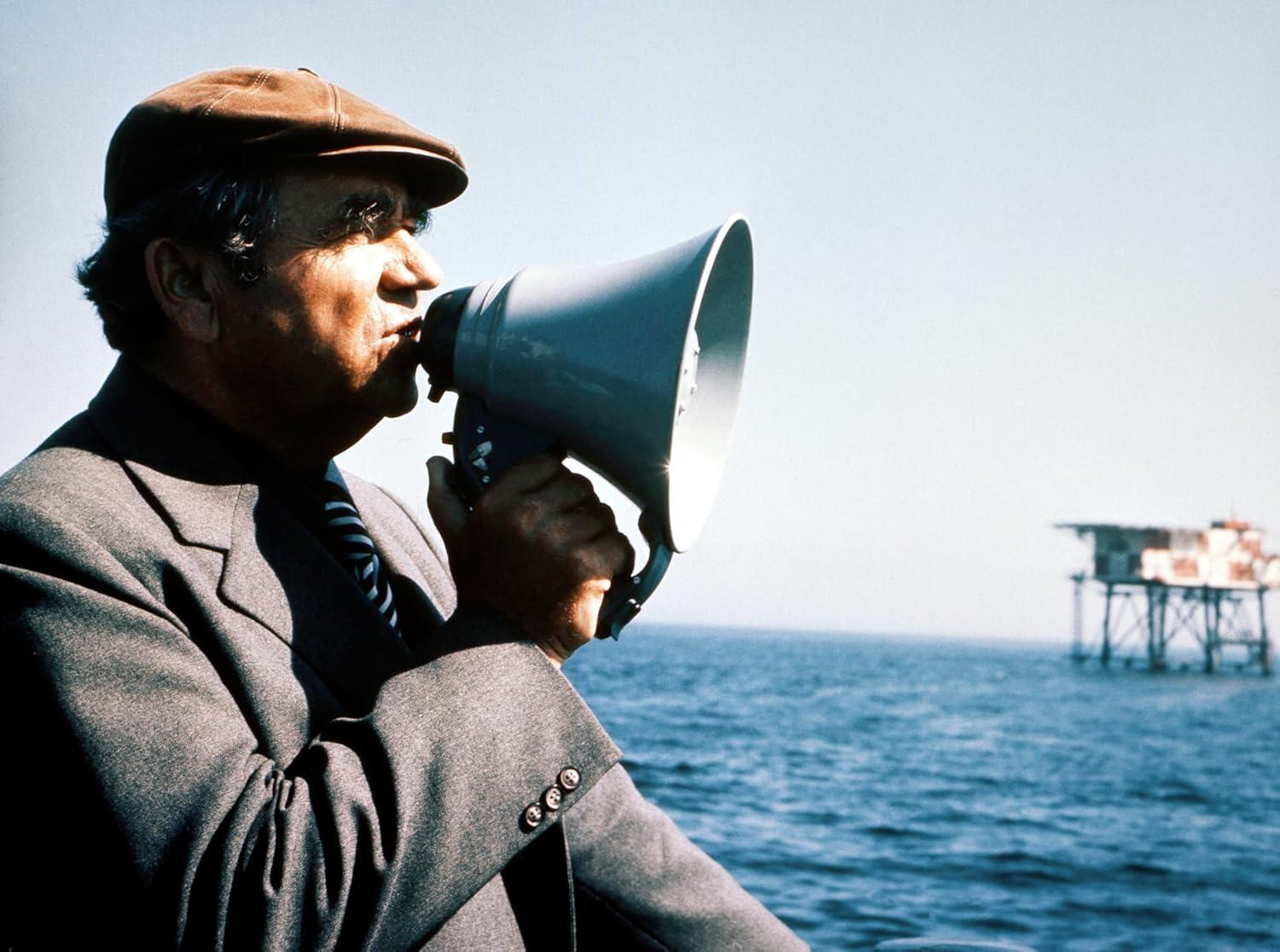 Ein Mann mit Hut spricht in ein Megaphon, mit Meer und Struktur im Hintergrund.
