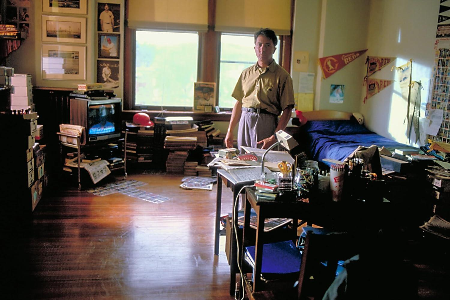 Ein unordentliches Zimmer mit einem stehenden Mann. Enthält Fernseher, Bücher und Sportmemorabilien.