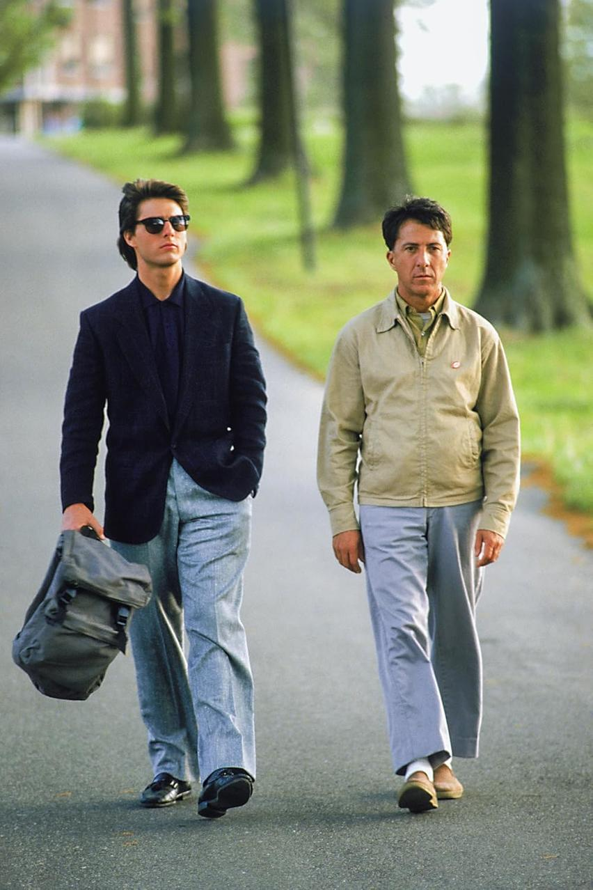 Zwei Männer gehen eine Straße entlang. Einer trägt Sonnenbrille und Jacke, hält eine Tasche. Der andere trägt eine beige Jacke.