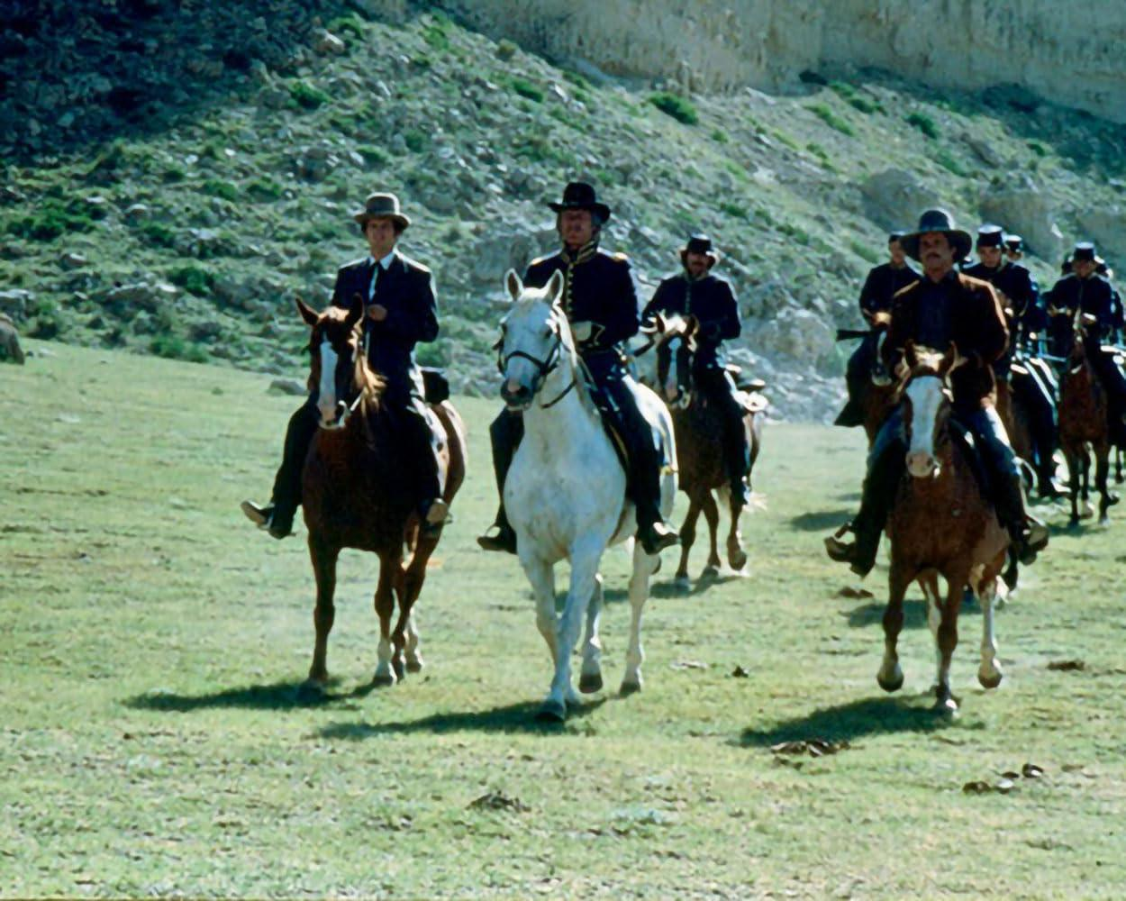 Eine Gruppe von Reitern auf Pferden auf einer grünen Wiese, mit Bergen im Hintergrund, tragen Anzüge.