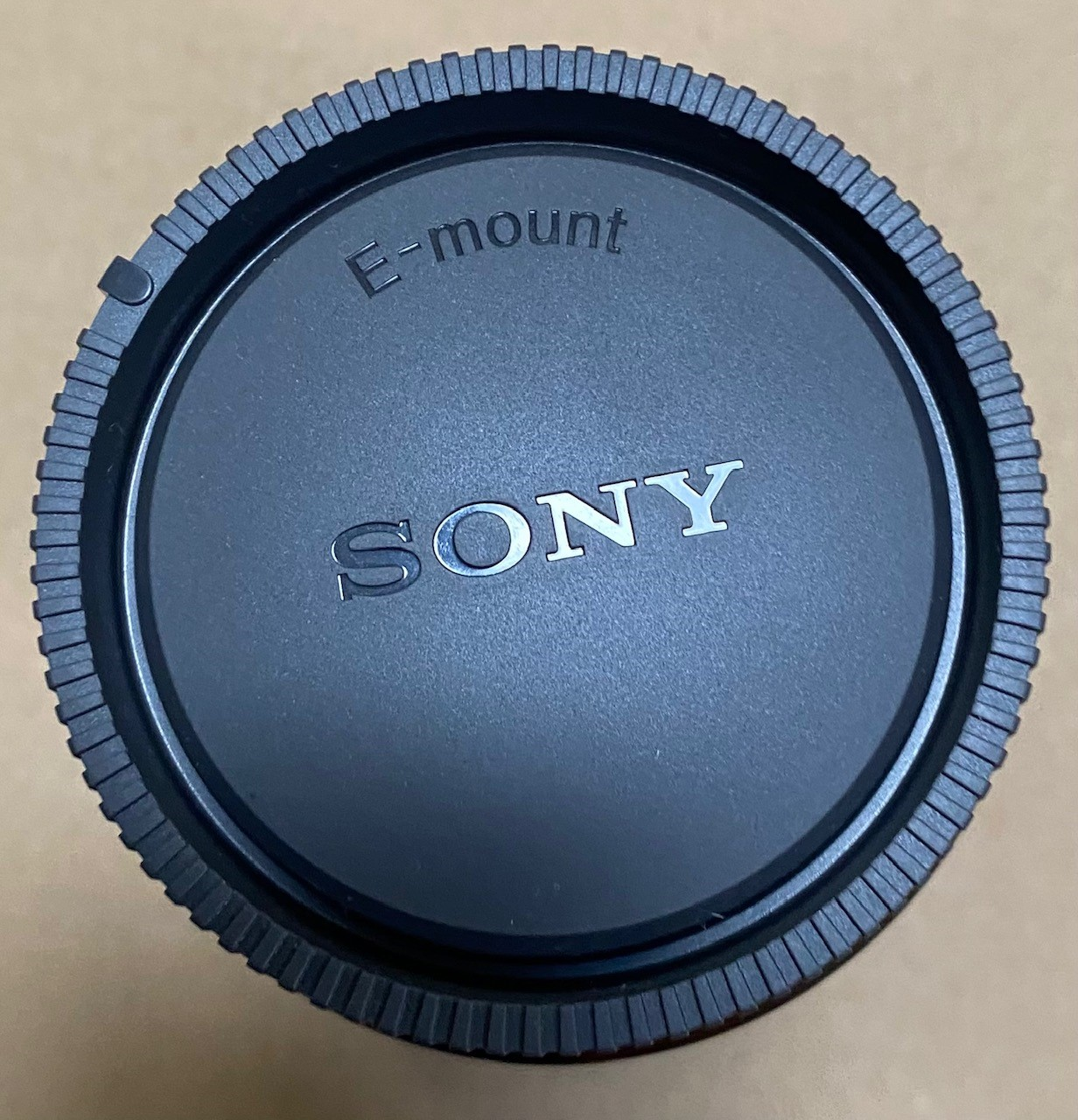 Czarna osłona obiektywu Sony z napisem "E-mount" i "SONY", na beżowej powierzchni.